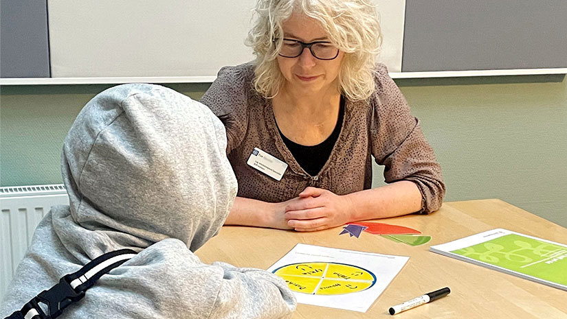 Eva Bjuvmar sitter vid ett bord mittemot ett barn. På bordet mellan dem ligger Mera koll på tid-material.